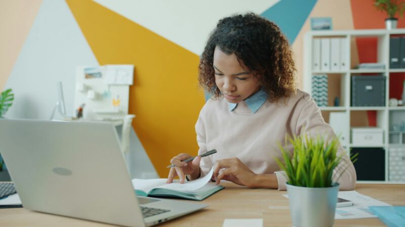 A woman writing in a notebook at a desk beside a laptop