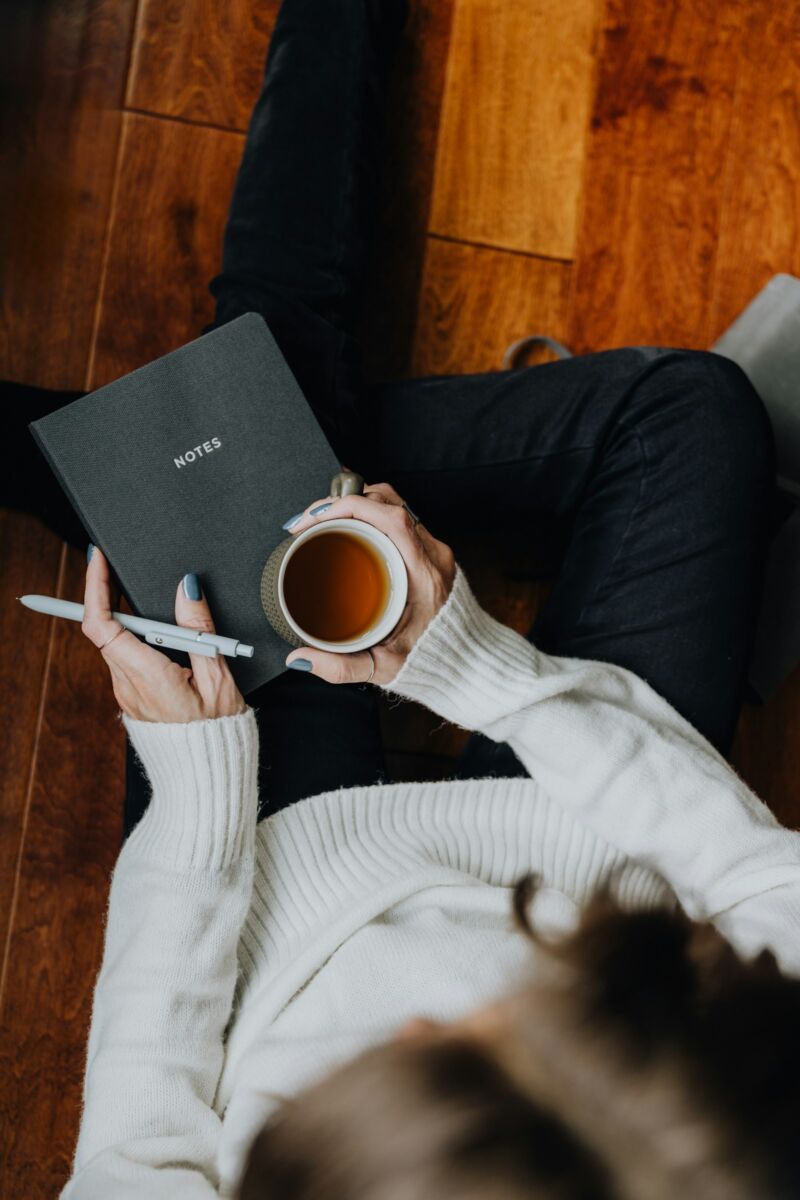 A woman in a cozy sweater holding coffee and a book